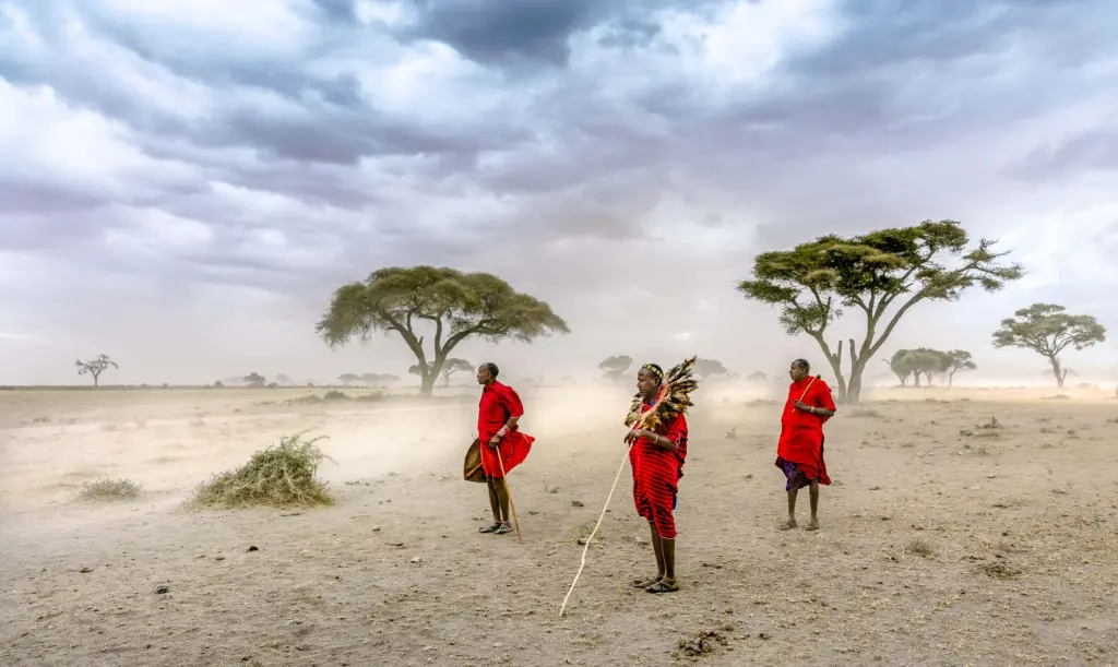 Drei Massai-Männer in traditionellen, leuchtend roten Tüchern (Shukas) stehen auf trockenem, staubigem Boden in einer weitläufigen afrikanischen Savanne. Die Luft ist durch einen starken Sandsturm gelblich-braun getrübt, der die Sicht auf vereinzelte, dunkelgrüne Akazienbäume im Hintergrund einschränkt. Der Himmel darüber ist bedeckt von schweren, dramatischen, dunkelgrauen Wolken. Die Männer halten Stäbe, einer trägt einen markanten Kopfschmuck aus Federn.